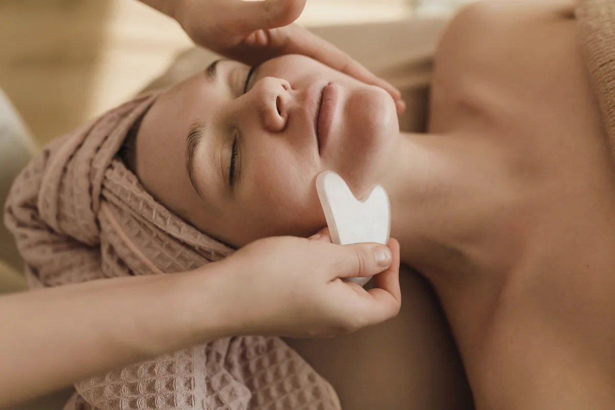 A relaxed client with a pink towel wrapped around her hair while a white heart-shaped stone is glided along her jaw