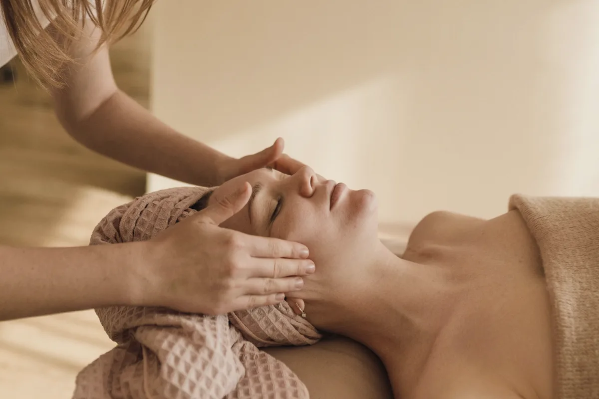 An esthetician's hands resting on the temples of a client lying on a facial bed, her hair wrapped in a pink towel