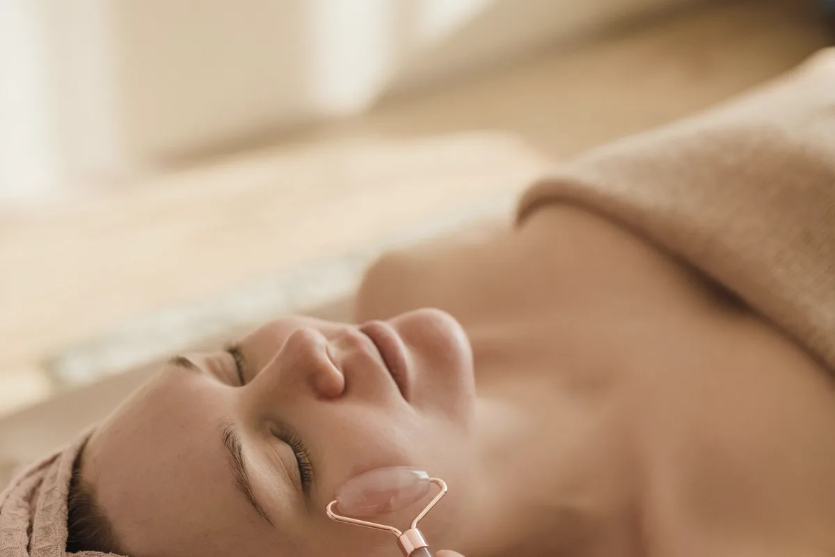 A woman relaxes with her eyes closed as a pink stone facial roller glides over her cheek.